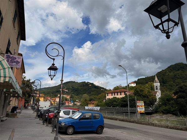 Via principale di Pedemonte con lampioni in ferro battuto, case colorate sulla collina e il campanile vicino al torrente