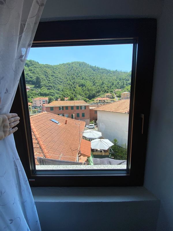 Vista dalla finestra: tetti in cotto di Pedemonte, casa con persiane verdi e colline coperte di boschi
